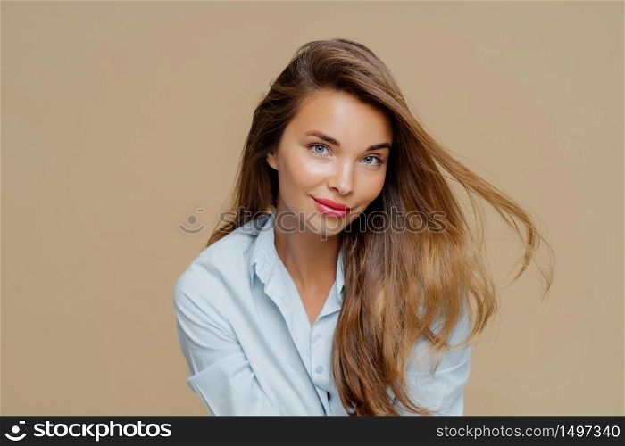 Portrait of attractive young woman has fair hair floating in wind, natural beauty, wears makeup, dressed in stylish shirt, isolated over brown background, has healthy skin. People, style, feminity