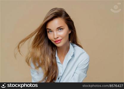 Portrait of attractive young woman has fair hair floating in wind, natural beauty, wears makeup, dressed in stylish shirt, isolated over brown background, has healthy skin. People, style, feminity
