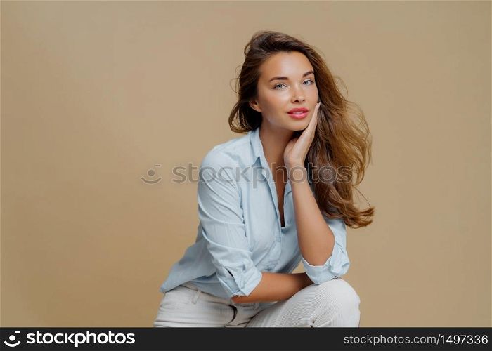 Portrait of attractive young woman sits against brown background, touches cheek, dressed in fashionbale outfit, has thoughtful look at camera, demonstrates her beauty. People and style concept