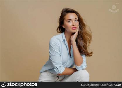 Portrait of attractive young woman sits against brown background, touches cheek, dressed in fashionbale outfit, has thoughtful look at camera, demonstrates her beauty. People and style concept
