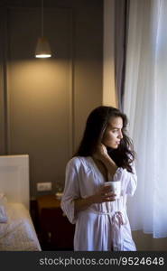 Portrait of attractive young woman with mug by the window in bedroom