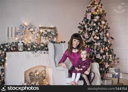 Portrait of baby and mother sitting on a chair near the Christmas tree.. Daughter and mom sitting in a chair near the Christmas tree 9631.
