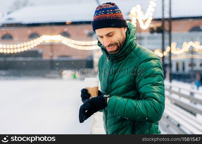 Portrait of bearded male dressed in warm clothes, looks at watch as waits for someone outdoor during frosty weather, drinks takeaway coffee. Handsome man waits for girlfriend, going to have date