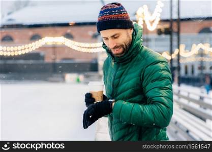 Portrait of bearded male dressed in warm clothes, looks at watch as waits for someone outdoor during frosty weather, drinks takeaway coffee. Handsome man waits for girlfriend, going to have date