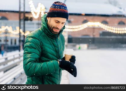 Portrait of bearded male dressed in warm clothes, looks at watch as waits for someone outdoor during frosty weather, drinks takeaway coffee. Handsome man waits for girlfriend, going to have date