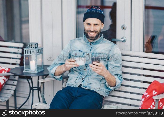 Portrait of bearded male with broad smile, sits at bench, uses modern cell phone, surfes social networks, happy to recieve good news, drinks aromatic beverage. People, technology, lifestyle concept