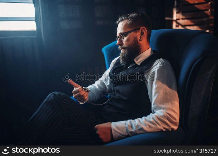 Portrait of bearded man in glasses sitting in armchair, looking at the pocket watch. Writer, journalist, literature author, blogger or poet concept. Man sitting in armchair, looking at pocket watch