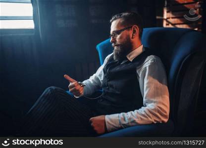 Portrait of bearded man in glasses sitting in armchair, looking at the pocket watch. Writer, journalist, literature author, blogger or poet concept. Man sitting in armchair, looking at pocket watch