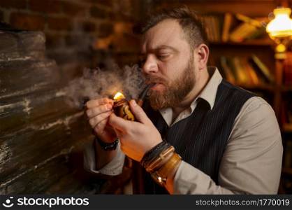 Portrait of bearded man lights smoking pipe with a match, closeup view. Tobacco smoking culture, specific rich flavor. Male smoker leisures in office. Bearded man lights smoking pipe with a match