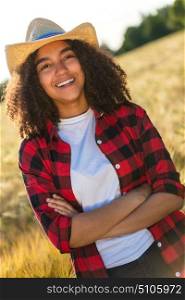 Portrait of beautiful and happy mixed race African American female girl child wearing straw cowboy hat and plaid shirt and white t-shirt, smiling with perfect teeth in a wheat or barley field at sunset