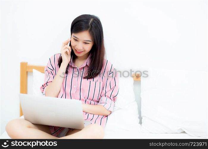 Portrait of beautiful asian young woman setting on bed using laptop computer and talk smart phone at bedroom for leisure and relax, freelance with girl working notebook and calling telephone, communication and lifestyle concept.