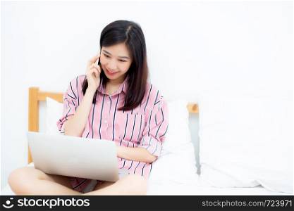 Portrait of beautiful asian young woman setting on bed using laptop computer and talk smart phone at bedroom for leisure and relax, freelance with girl working notebook and calling telephone, communication and lifestyle concept.
