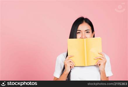Portrait of beautiful Asian young woman teen smile covering her face with yellow book, female person hiding behind an open book show only eyes, studio shot isolated on pink background