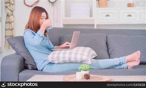 Portrait of beautiful attractive Asian woman using computer or laptop holding a warm cup of coffee or tea while lying on the sofa when relax in living room at home. Lifestyle women at home concept.