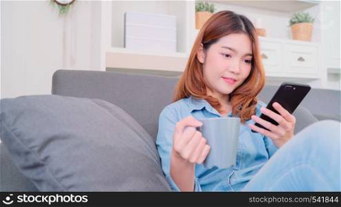 Portrait of beautiful attractive smiling Asian woman using smartphone holding a warm cup of coffee or tea while lying on the sofa when relax in living room at home. Lifestyle women at home concept.