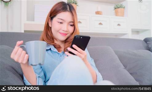 Portrait of beautiful attractive smiling Asian woman using smartphone holding a warm cup of coffee or tea while lying on the sofa when relax in living room at home. Lifestyle women at home concept.