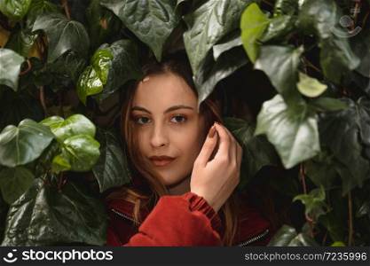 Portrait of beautiful attractive young girl stands among green leaves and looks at camera