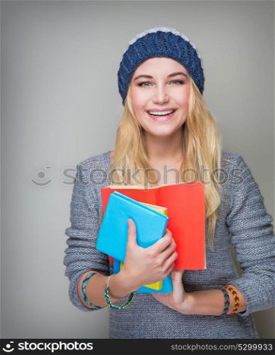 Portrait of beautiful blond student girl wearing stylish hat and holding in hands colorful books standing over gray background