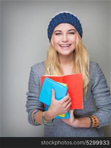 Portrait of beautiful blond student girl wearing stylish hat and holding in hands colorful books standing over gray background