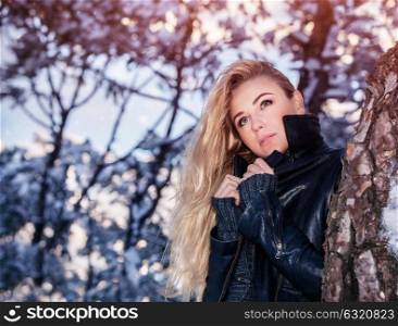 Portrait of beautiful blond woman standing near tree in the winter park, fashionable clothing, gorgeous model posing outdoors