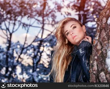 Portrait of beautiful blond woman standing near tree in the winter park, fashionable clothing, gorgeous model posing outdoors
