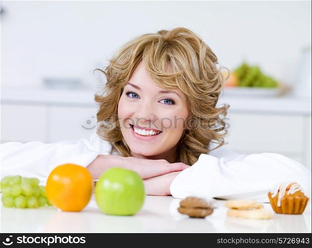Portrait of beautiful blond woman with fruits and cakes sitting on the kitchen