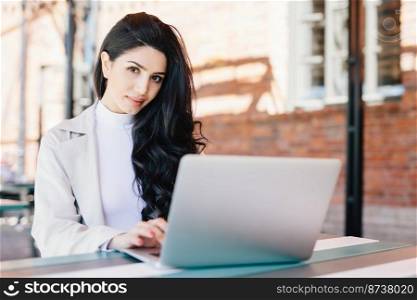 Portrait of beautiful brunette female freelancer typing a message via email using laptop while sitting on terrace looking at camera with confident look. Young lady working with computer outdoors