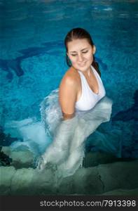 Portrait of beautiful brunette woman in long white dress posing in swimming pool