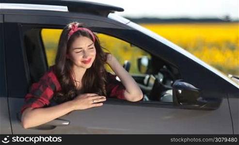Portrait of beautiful brunette woman leaning out of car window, enjoying amazing landscape during summer roadtrip in countryside. Relaxed happy girl with long hair looking out of automobile window and smiling on sunny day during travel vacation.