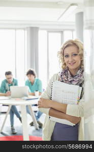 Portrait of beautiful businesswoman holding files with colleagues working in background at creative office