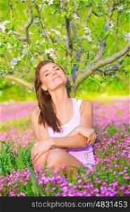 Portrait of beautiful calm girl with closed eyes sitting down on pink floral glade under blooming apple tree, relaxation outdoors in spring time