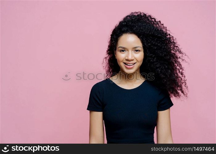 Portrait of beautiful charming dark skinned woman smiles happily, pleased to hear pleasant news, wears casual black t shirt, models against rosy background, copy space aside for your promotion