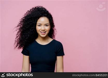 Portrait of beautiful charming dark skinned woman smiles happily, pleased to hear pleasant news, wears casual black t shirt, models against rosy background, copy space aside for your promotion