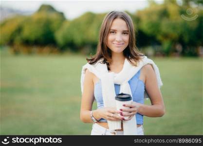 Portrait of beautiful charming woman with short dark hair, has healthy skin, dressed in casual clothes, holds takeaway coffee, enjoys recreation time, stands against blurred nature background