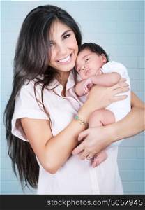 Portrait of beautiful cheerful mother with cute little baby on hands over blue wall background, enjoying parenthood, love and happiness concept