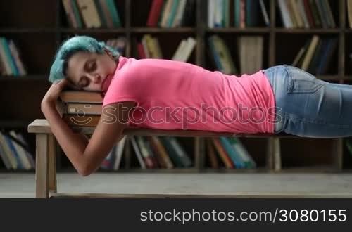 Portrait of beautiful college female who fell asleep on a stack of books in library. Closeup. Exhausted hipster girl with blue hair napping on pile of books while preparing for exams in university library. Education, exams concept.