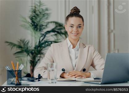 Portrait of beautiful confident business lady in elegant clothes smiling pleasantly while sitting at workplace in office and using modern technologies, working on laptop. Job and occupation concept. Cheerful business lady in elegant clothes smiling pleasantly at camera