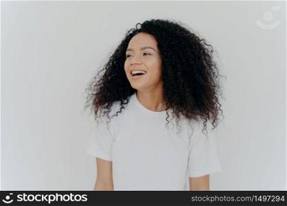 Portrait of beautiful curly adult woman turns gaze aside, has glad expression, smiles and laughs at something funny, dressed in white t shirt, models indoor, feels free, energized and relieved,