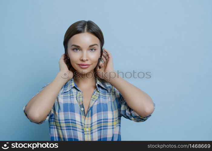 Portrait of beautiful dark haired European woman has healthy glowing skin, make up, dressed in casual checkered shirt, looks directly at camera, stands against blue background, being in good mood
