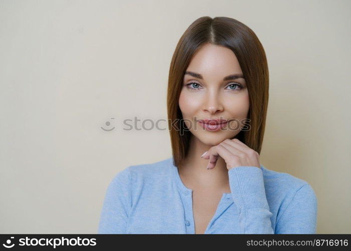 Portrait of beautiful dark haired woman with healthy natural clean skin, touches chin, has manicure and wears blue jumper, isolated over beige background. People, beauty, face expressions concept.