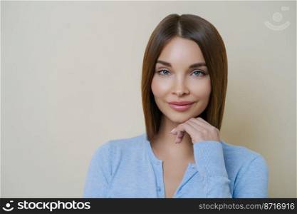 Portrait of beautiful dark haired woman with healthy natural clean skin, touches chin, has manicure and wears blue jumper, isolated over beige background. People, beauty, face expressions concept.