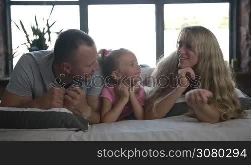 Portrait of beautiful family lying on bed. Little curly blonde daughter with ponytail looking at camera and smiling while her parents kissing her cheeks. Loving parents kissing thier little girl while relaxing on bed at home. Slo mo. Dolly shot.