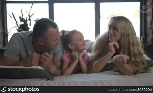 Portrait of beautiful family lying on bed. Little curly blonde daughter with ponytail looking at camera and smiling while her parents kissing her cheeks. Loving parents kissing thier little girl while relaxing on bed at home. Slo mo. Dolly shot.