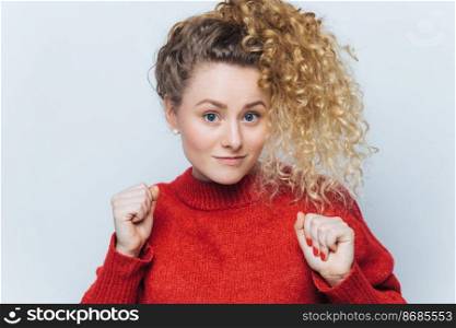 Portrait of beautiful female with curly hair, raises fists, dressed in casual red sweater, isolated over white background, poses in studio. People, beauty, facial expressions and body language concept