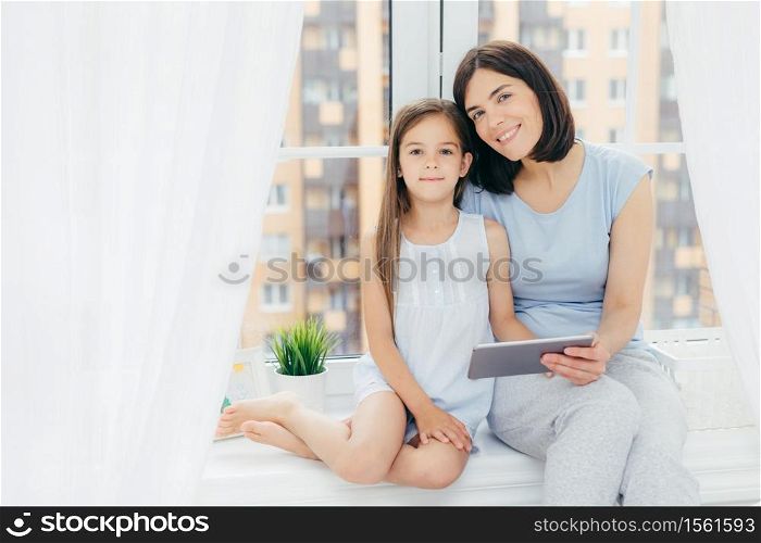 Portrait of beautiful female with dark hair, dressed in casual clothes, holds digital tablet, sits near her daughter, watch something, sit on window sill, play in bedroom. People, lifestyle and family