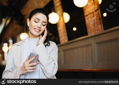 Portrait of beautiful female with dark hair tied in pony tail dressed formally listening to music with earphones closing her eyes with relaxation holding cell phone. People, lifestyle, technology