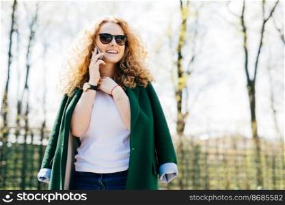 Portrait of beautiful female with fluffy blonde hair talking on cell phone to her friend, looking happily and excited standing against green park background holding hand on neck. Human emotions