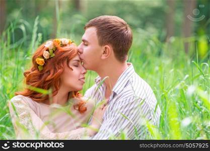 Portrait of beautiful gentle loving couple sitting on the grass in forest, romantic guy with pleasure kissing his cute girlfriend, enjoying romantic relationship