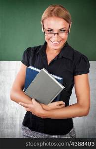 Portrait of beautiful happy young teacher standing in the classroom near chalkboard with books in hands, back to school