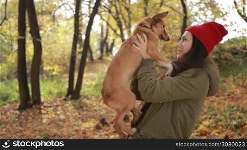 Portrait of beautiful hipster girl in stylish outfit holding her cute puppy and smiling in public park over gold autumn background. Attractive brunette woman and her dog enjoying good autumn weather in indian summer. Slow motion.